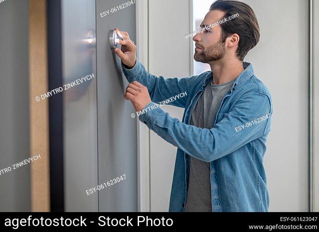 Examination. Young bearded man standing sideways to camera looking attentively touching device on wall indoors