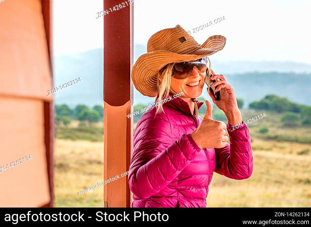 Happy woman standing on porch of rural ranch. She is using a mobile phone, smiling with thumbs up gesture, RU OK, lifeline, success, etc