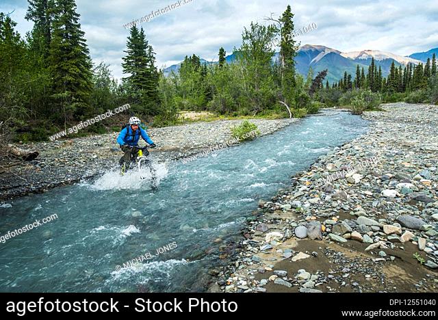 A man fat biking across a stream in Wrangell-St. Elias National Park and Preserve on a cloudy summer day in South-central Alaska; Alaska