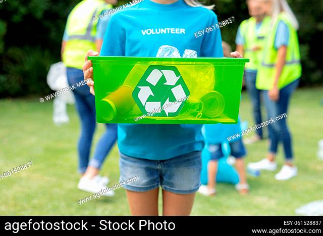 Caucasian girl holding recycling box with group of men and women collecting rubbish in field behind