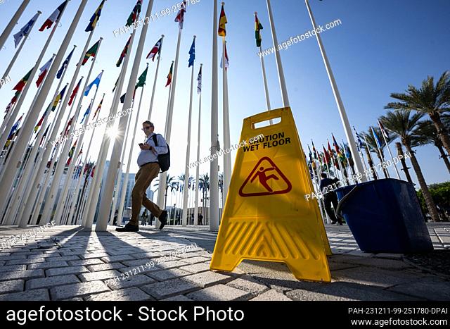 11 December 2023, United Arab Emirates, Dubai: A man walks in front of a sign reading ""Caution Wet Floor"" on the grounds of the UN Climate Change Conference...