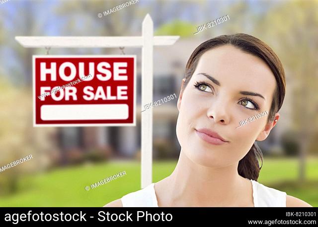 Thoughtful pretty mixed-race woman in front of home and house for sale real estate sign looking up and to the side