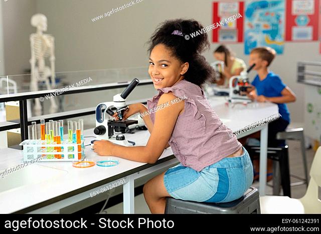 Portrait of smiling biracial elementary schoolgirl with microscope sitting at desk in laboratory