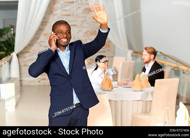 Handsome businessman in navy blue suit waving to someone and speaking over mobile phone while his foreign collegues sitting at table and communicating
