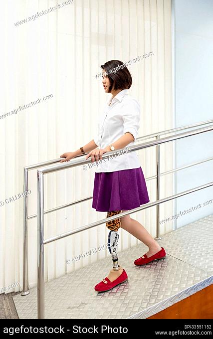 Young woman with leg prosthesis in therapy, walking on a ramp with handrails; Bangkok, Thailand