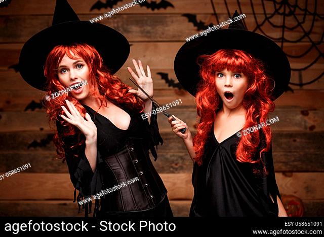 Halloween Concept - Beautiful caucasian mother and her daughter with long red hair in witch costumes and magic wand celebrating Halloween posing with over bats...