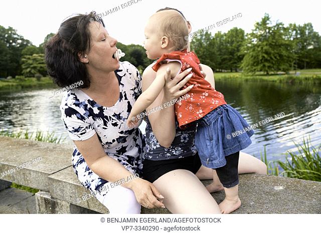 humorous grandmother with pacifier in mouth tasing baby toddler granddaughter, outdoors in park at lakeside