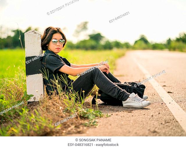 Young asian woman short hair and wearing sunglasses sit with backpack hitchhiking along a road wait for help in country road Thailand