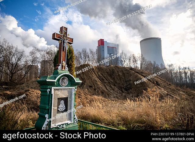07 January 2023, Poland, Bogatynia: A cross stands by the wayside in the Trzciniec district. In the background is the Turów large-scale lignite-fired power...