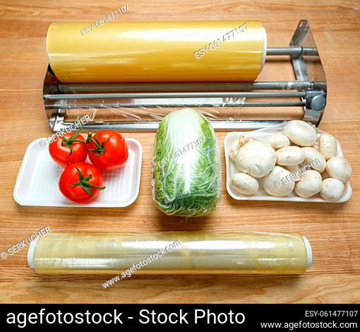 Rolls of transparent polyethylene food film for packing food on table with vegetables. Tomatoes, mushrooms and Chinese cabbage are wrapped in cling film for...