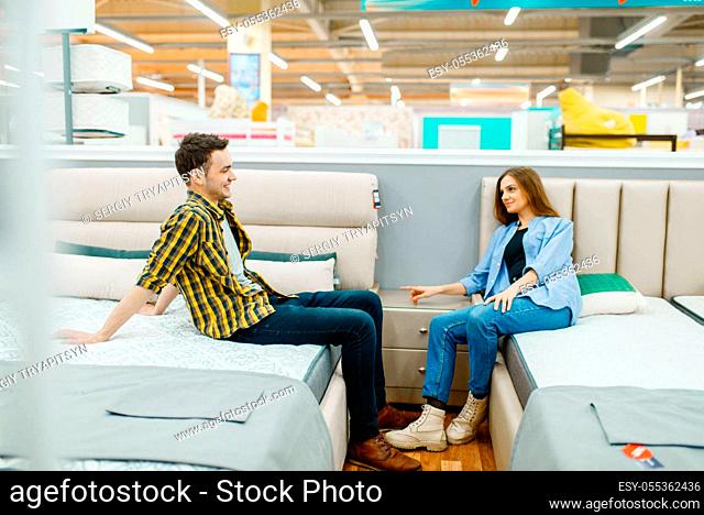 Young love couple sitting on beds in furniture store. Man and woman looking samples for bedroom in shop, husband and wife buys goods for modern home interior