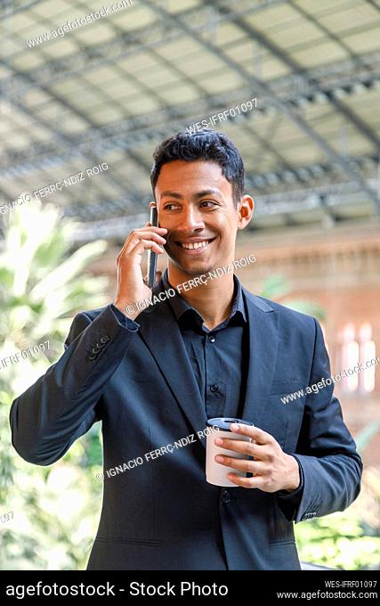 Happy young businessman holding disposable coffee cup talking on mobile phone