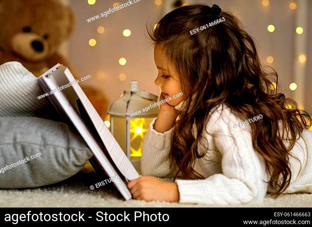 Happy little child girl is reading fairy tale book on the background with lights. Merry Christmas