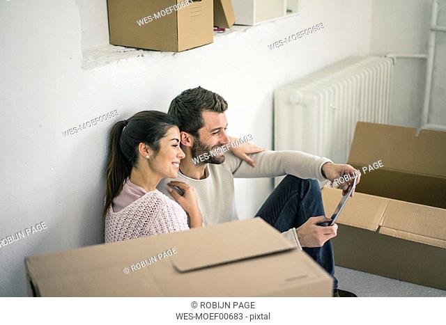Couple sitting in new home surrounded by cardboard boxes looking at tablet