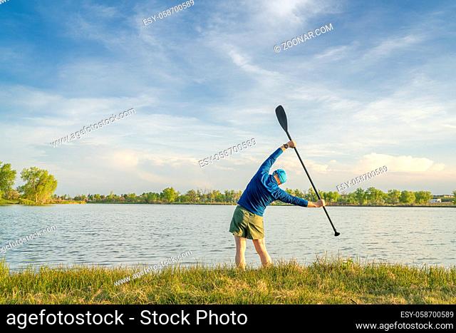 mature stand up paddler is stretching and warming up on a lake shore before paddling workout