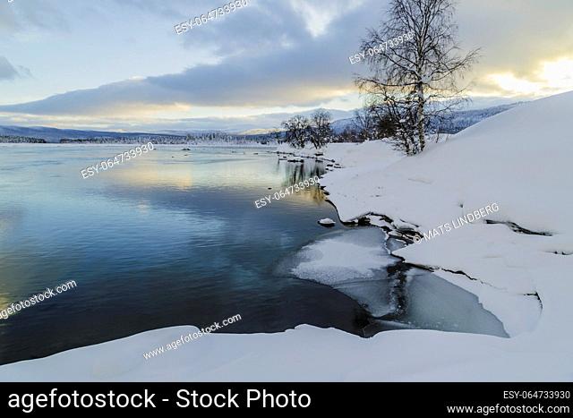 Winter landscape in Tjåmotis with mountains in background, sun shining over the mountains, creek with open water, sky with nice colors reflecting in the water