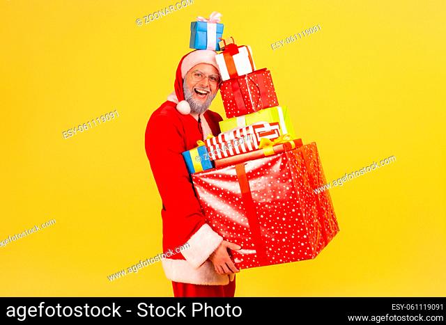 Happy funny elderly man with gray beard in santa claus costume holding stack of Christmas presents in hands and on head, celebrating with new year