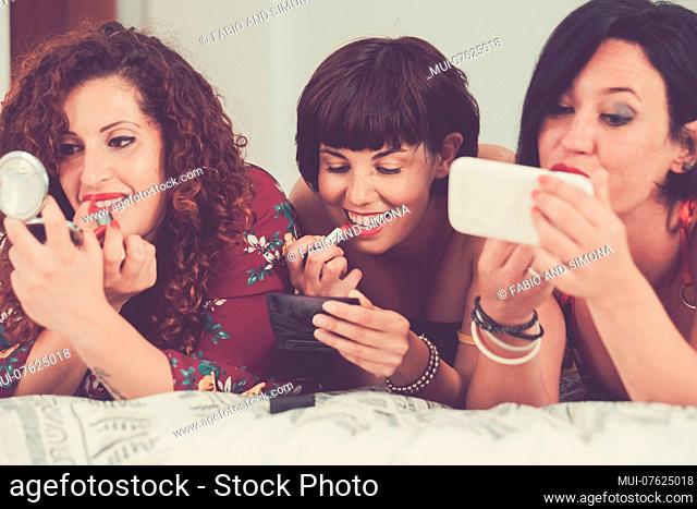 three females friends stay together lay down on the bed doing make up before go out to have party night. friendship concept one of them use a mobile phone like...