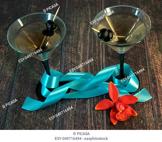 Two martini glasses with olives, with blue ribbon, red orchid and gift box on a wooden table. Close-up