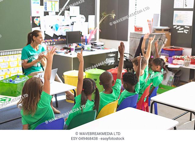 Front view of caucasian teacher discussing about green energy and recycle while multi ethnic students raising their hands at desk in classroom