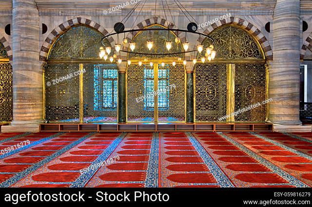 Interior shot of three arched ornate engraved golden doors, big chandelier over marble wall with pillars and red decorated carpet at Suleymaniye Mosque