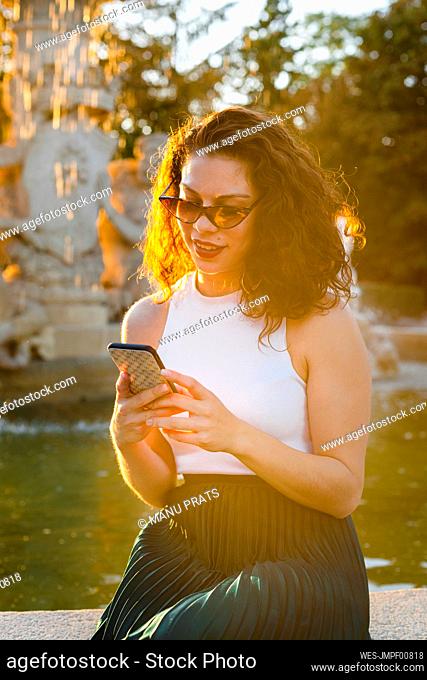 Woman wearing sunglasses using smart phone while sitting against fountain in park