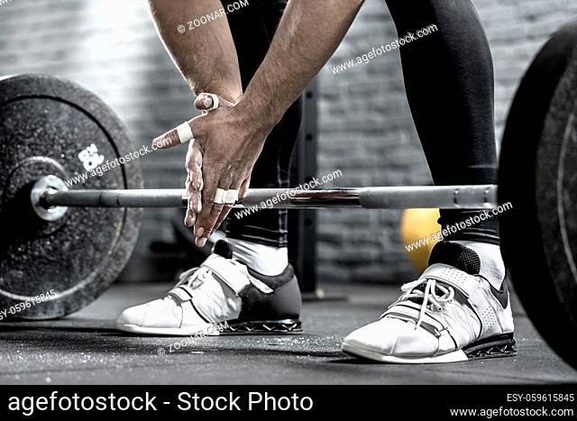 Picture of man's hands in front of the barbell on the background of his legs in black pants and white sneakers. Guy holds the hands together