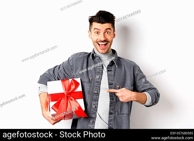 Happy Valentines day. Excited caucasian guy pointing at gift box from lover, smiling and looking amazed, buying presents on romantic date, white background