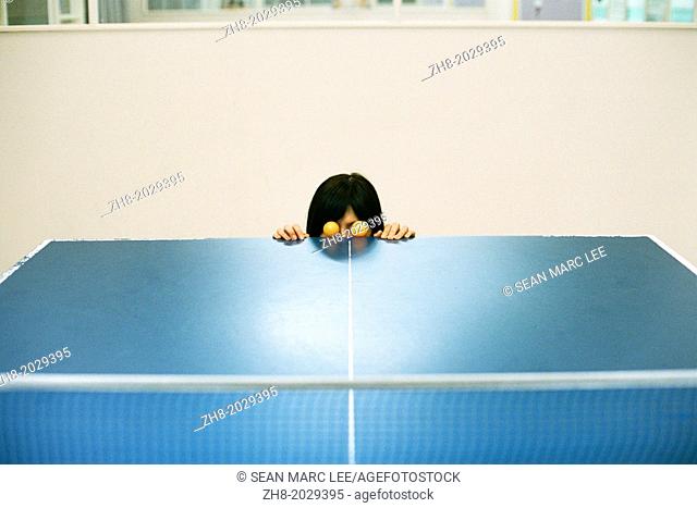 A woman poses with two orange ping pong balls as eyes at the edge of a ping pong table