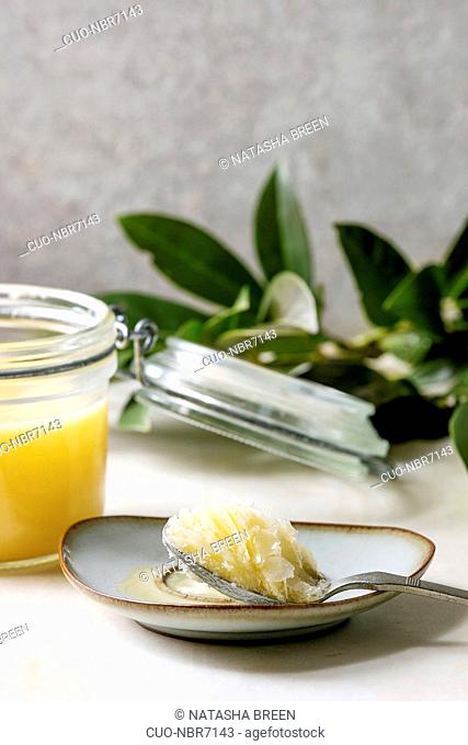 Homemade Melted ghee clarified butter in open glass jar and spoon on saucer standing on white marble table with green branch. Grey wall at background