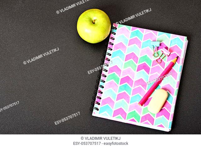 Top view of girly pink notebook with pencil, eraser, binder clips and apple on black background