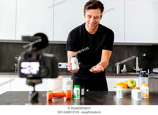 Cheerful young man filming his video blog episode about healthy food additives while standing at the kitchen table and showing pills