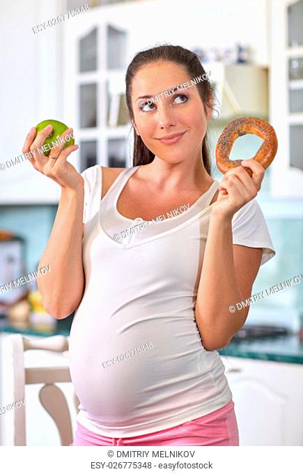 Young pregnant woman with apple and bagel in her hands in the house kitchen. Concept of healthy food