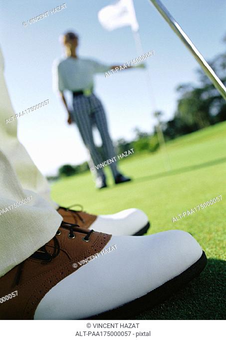 Golfer's feet on green, and second golfer holding pole