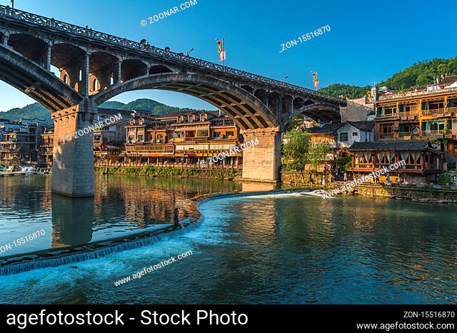 Feng Huang, China - August 2019 : Small cascade under the road bridge over Tuo Jiang river and wooden houses in ancient old town of Fenghuang known as Phoenix