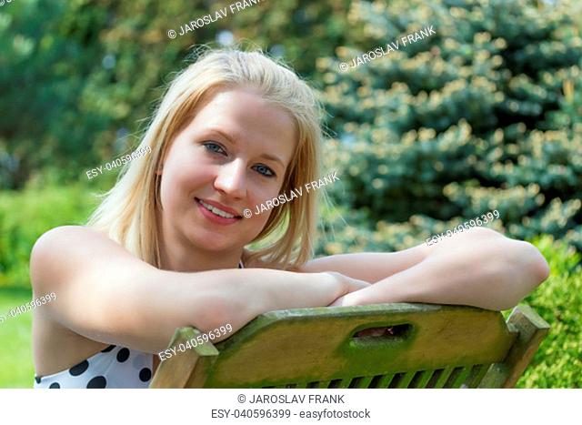 Closeup portrait of cool smiling young blond woman sitting on the wooden chair in the garden. Woman is looking at the camera