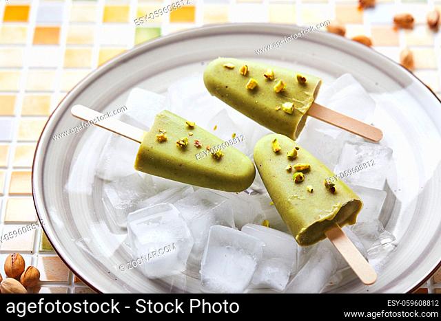 Frozen homemade pistachio popsicle in bowl of ice on mosaic tile table. Refreshing popsicle, frozen green juice on stick. Top view, copy space