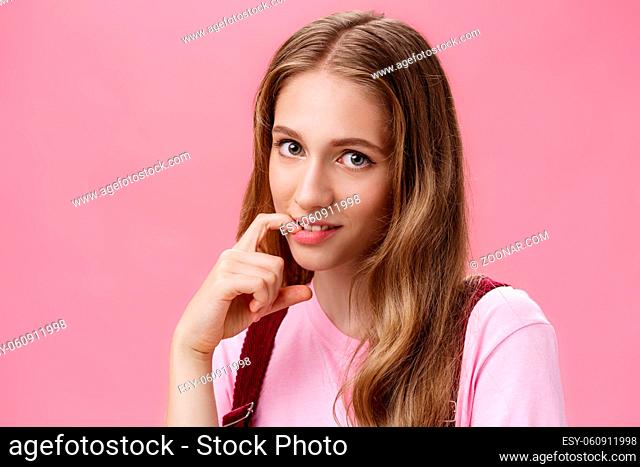 Cute feminine girl learning how make glances and seduce boyfriend with flirty gaze biting finger sensually posing against pink background