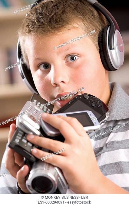 Young boy wearing headphones in bedroom holding many electronic devices
