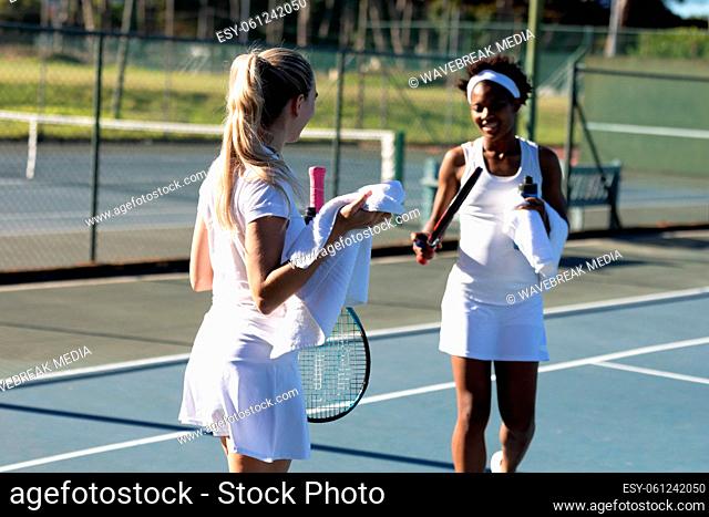 Smiling female multiracial players with towel talking during break at tennis court on sunny day
