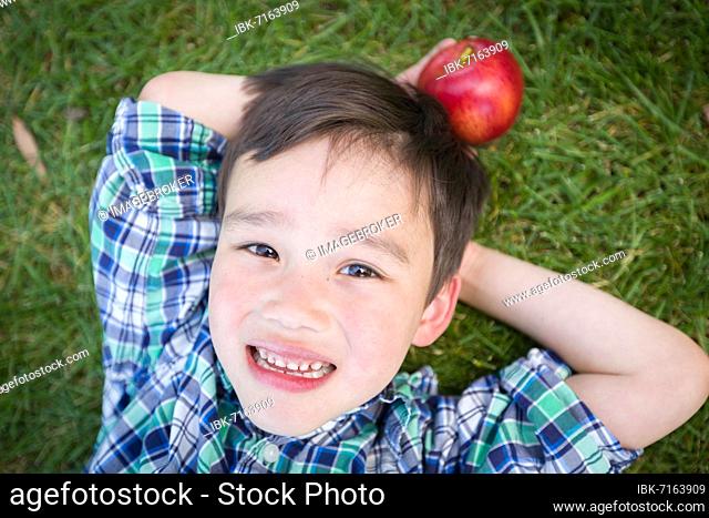 mixed-race chinese and caucasian young boy with apple relaxing on his back outside on the grass