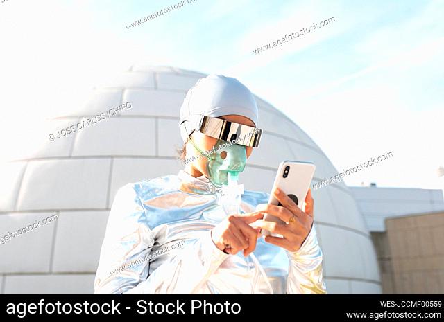 Woman in protective suit and oxygen mask using mobile phone while standing against igloo