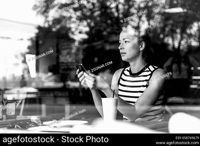 Thoughtful caucasian woman holding mobile phone while looking through the coffee shop window during coffee break. Street reflections in the window glass