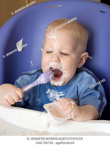A baby makes a big mess as he sits in his high chair feeding himself strawberry yoghurt