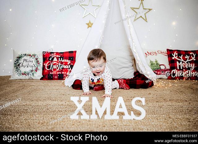 Smiling baby girl crawling toward XMAS text on floor at home during Christmas