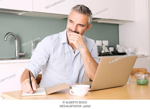 Smiling man writing in notebook and using laptop in home office