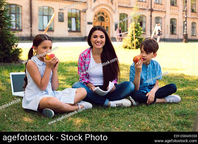 Children Sitting On School Yard And Eating Apples. Teacher Sitting Nearby And Smiling. Relaxing Lesson Concept