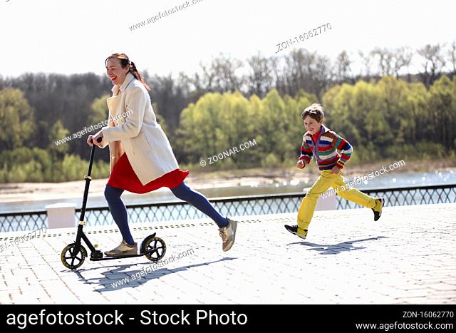 A woman is riding a scooter and her son runs after her. Mom and son on a walk in the park