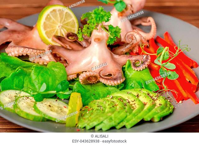 close up of octopus served with sliced avocado, lettuce, red pepper, lime, cucumber and sprig of pea leaves on plate, natural seafood