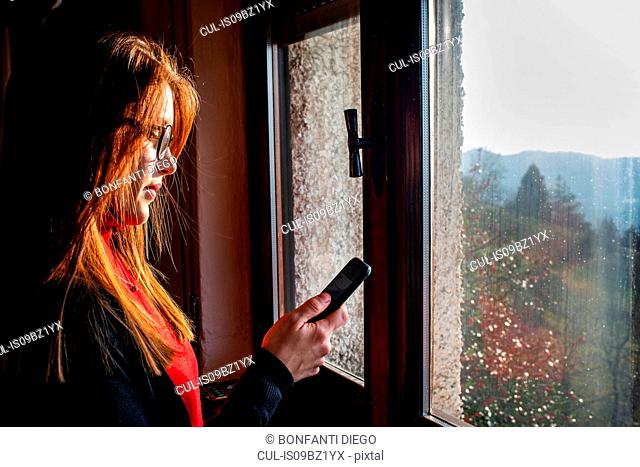 Young woman with long red hair looking at smartphone by window